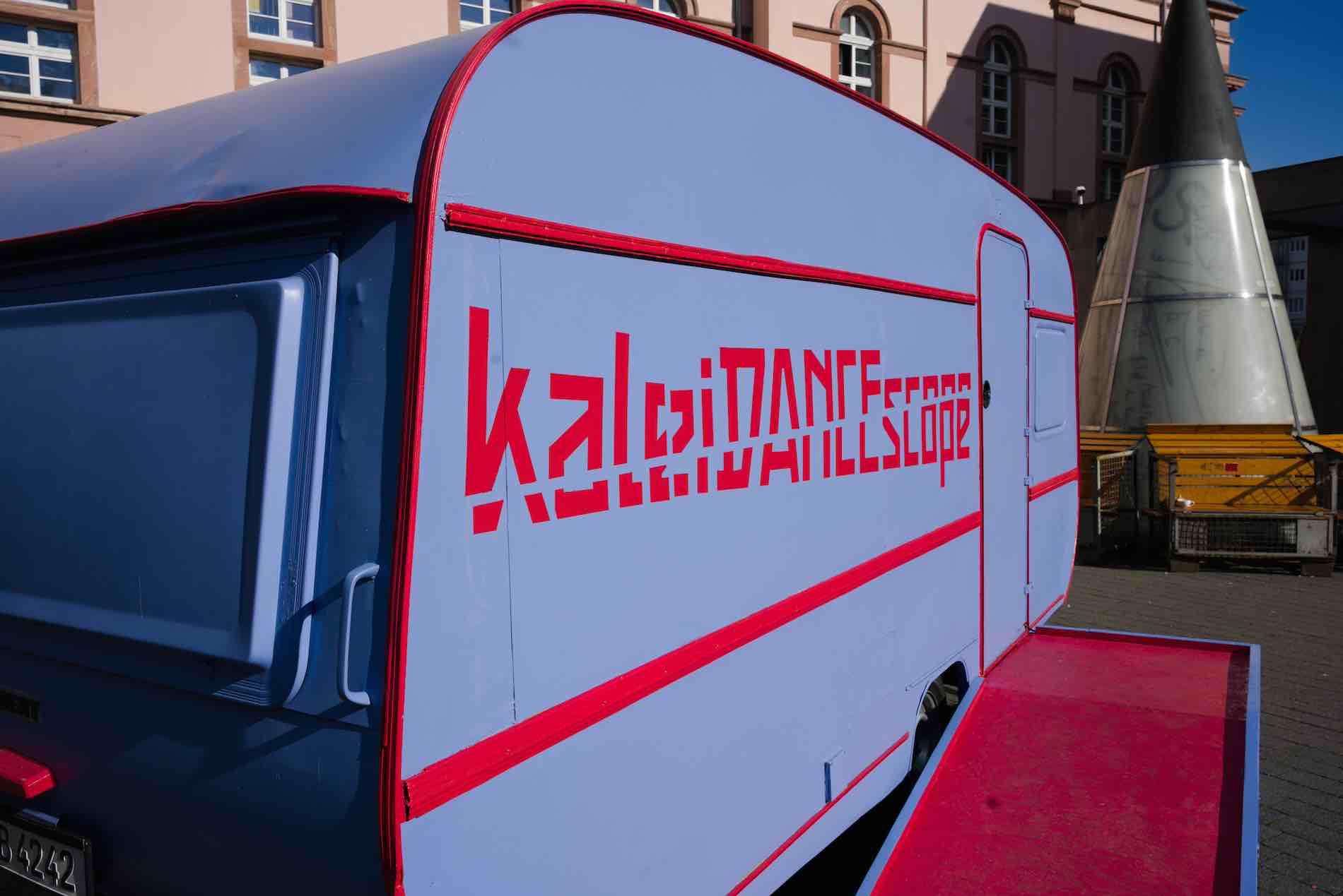 The kaleiDANCEscope lettering on the blue and red caravan can be seen in the bright sunlight. In the background is a silver cylindrical sculpture.
photo: Cornelius Schaper