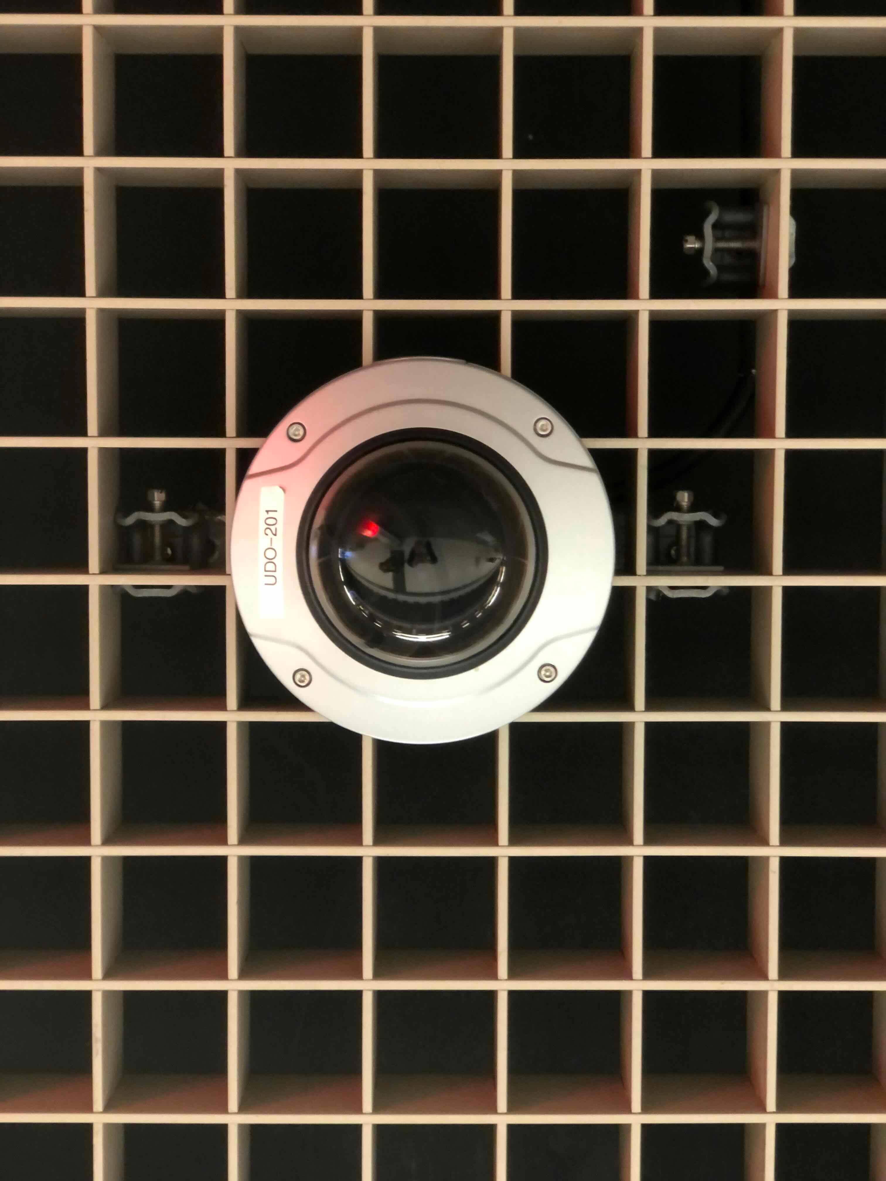 A round surveillance camera mounted on a metal grille. The camera has a small red light and a label that reads 'UDO-201'.