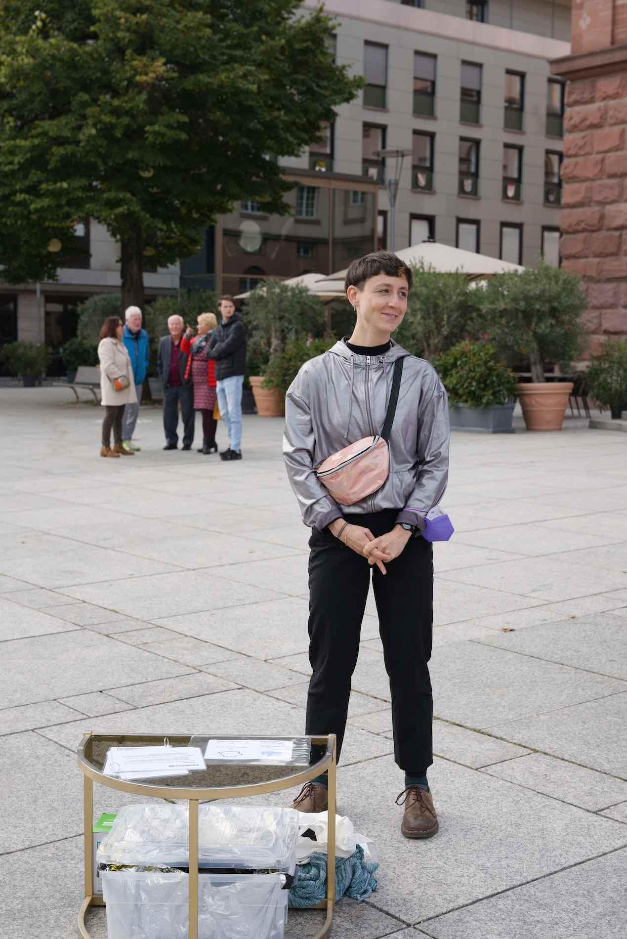 Performer Mia Werner stands in the square. Mia wears a silver jacket and smiles in the direction of potential visitors.
photo: Cornelius Schaper