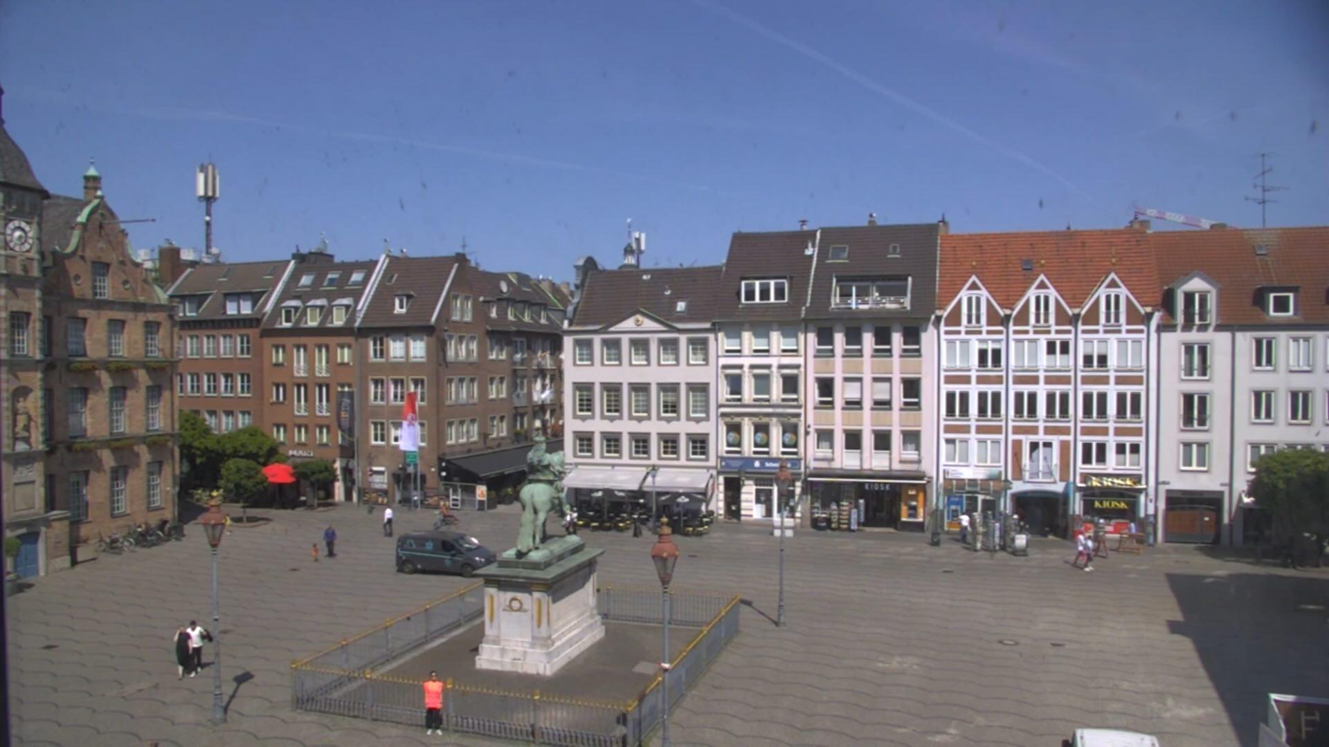 A large, open square in a city with old and new houses. In the middle stands a statue on a pedestal. Some people are walking around the square, and there are shops on the ground floor of the houses. In the foreground, a person in an orange T-shirt is looking at the camera. The sky is blue and sunny.