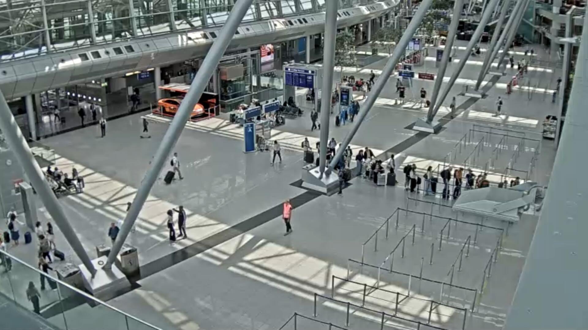 Perspective of a surveillance camera. A lot of people a visible in a huge, bright entrance hall of an airport. There are high ceilings, large windows, signs, luggage, and security checkpoints. In the foreground, there are lines at security checks. In the background, passengers and an orange airport bus are visible. In the center is one person in an orange west located.
