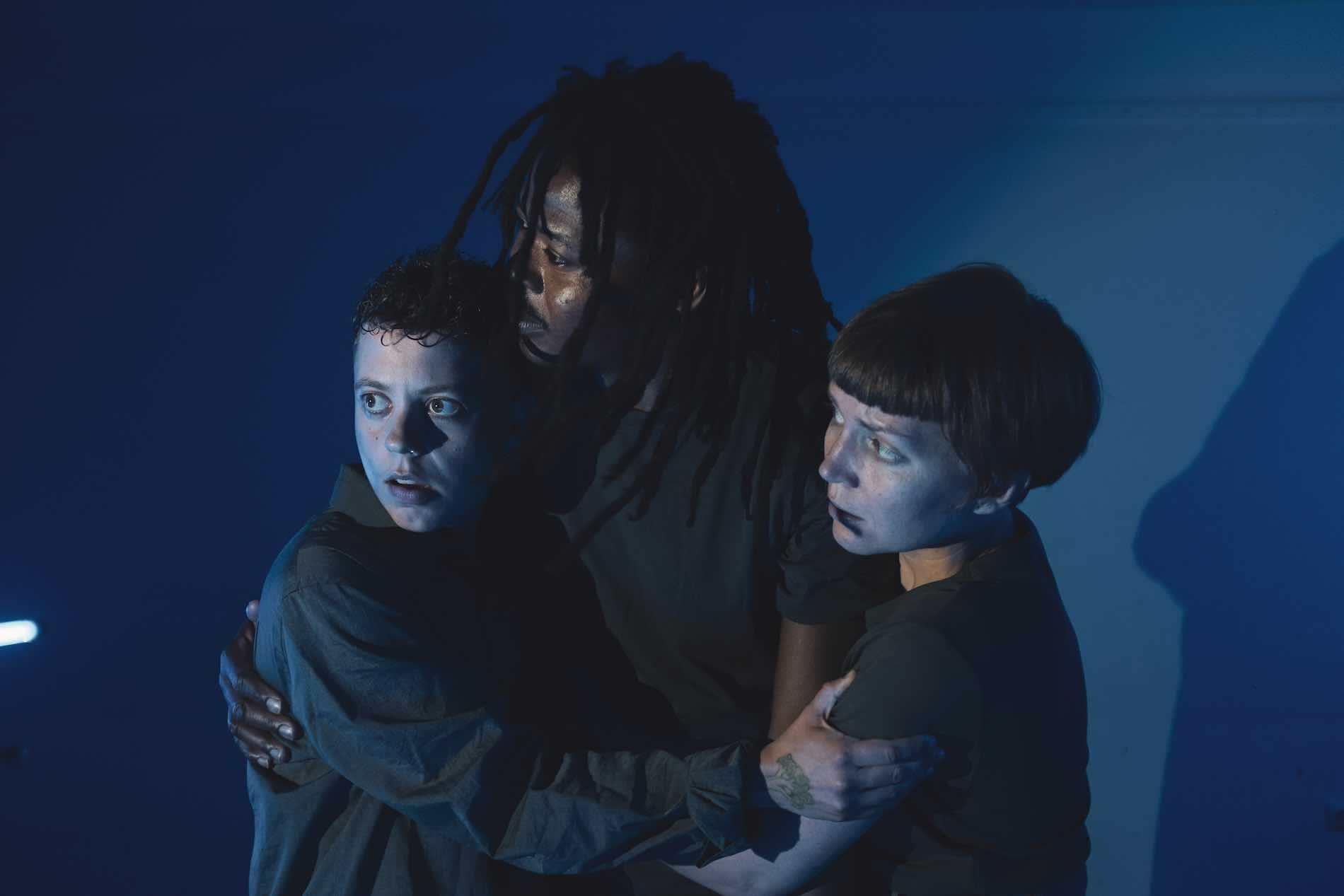 3 people hold on to each other in a half-embrace. The blue light draws attention to their frightened faces.