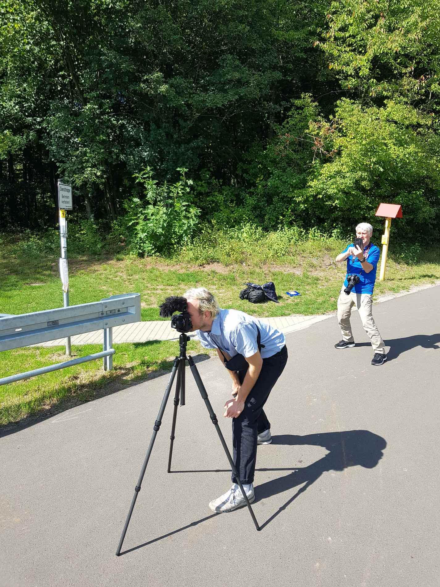 A person is standing behind a tripod with a camera. Behind them is another older person taking photographs.