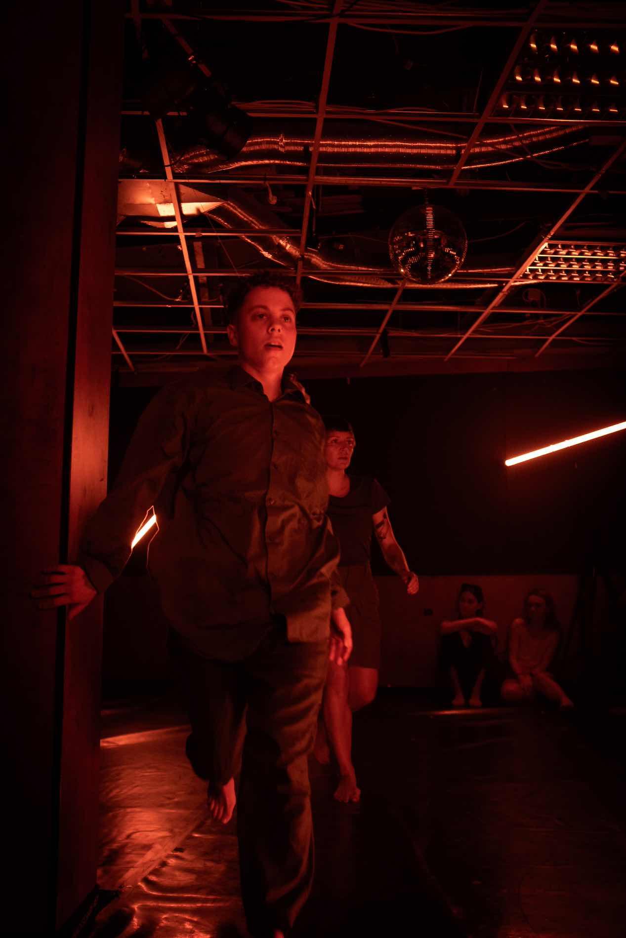 Two people run away in a room bathed in red light. In the background, an audience sits on the floor.