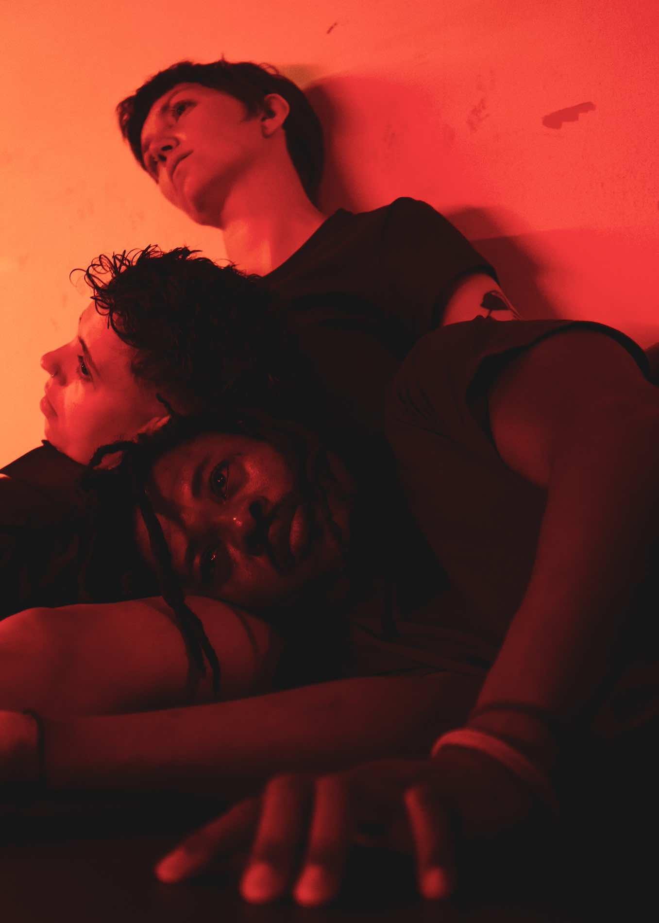 In red light, three people lie motionless on top of each other with their eyes open.