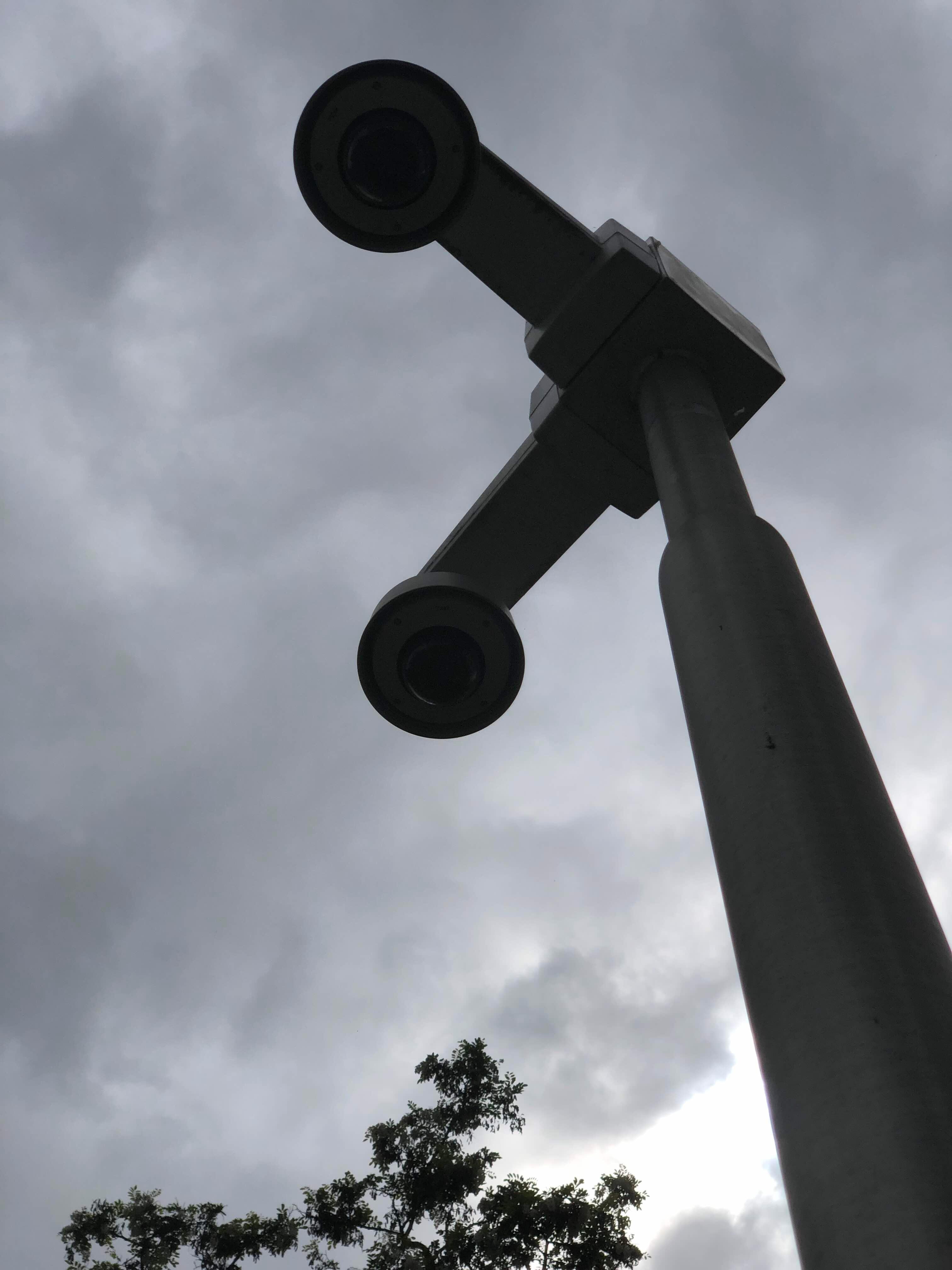 Large black sculpture with two round elements, resembling an oversized pair of binoculars or a camera pointing at the sky. Clouds and trees are visible in the background.