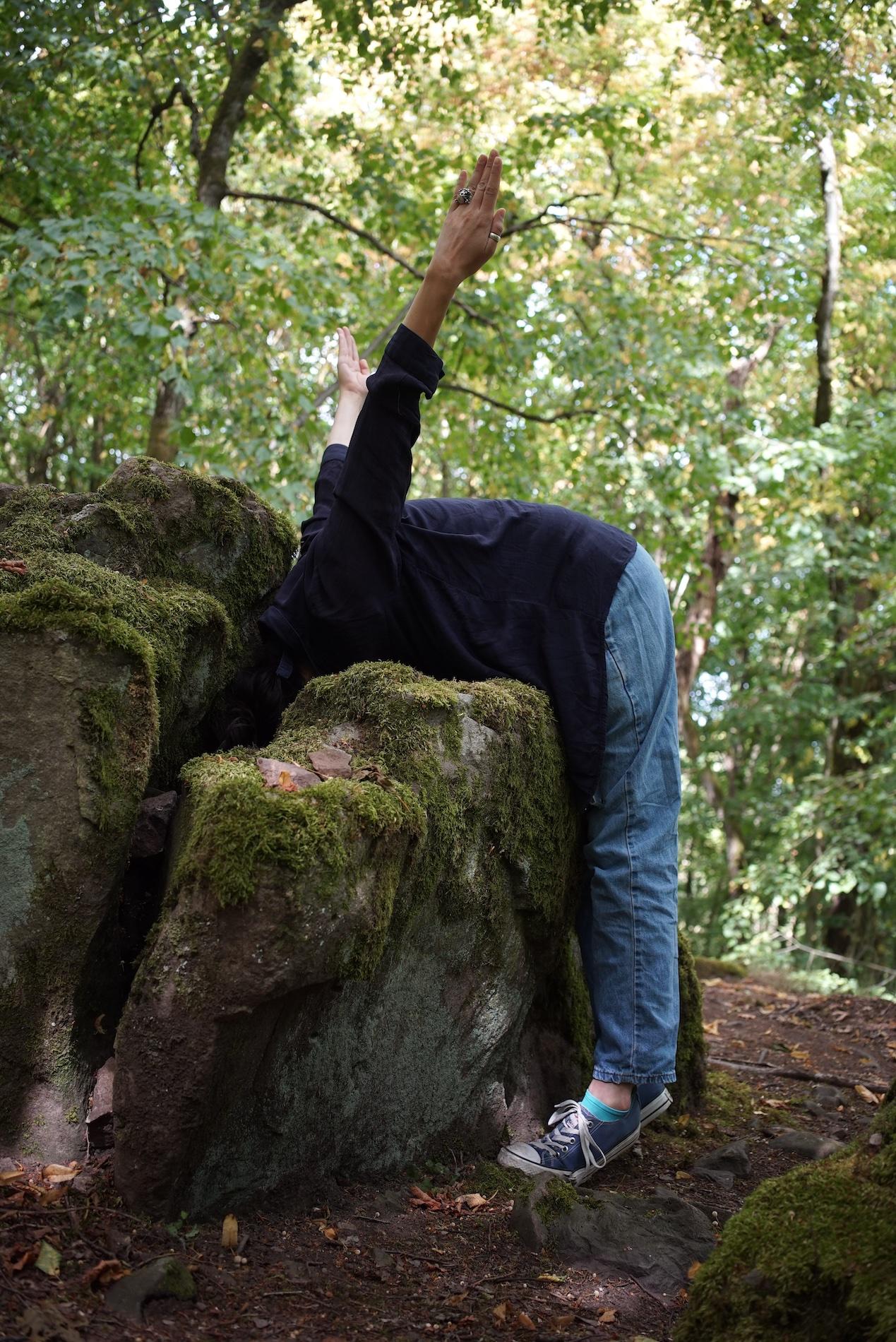 A performer is bent over a large stone in the forest and integrates the body into the natural environment.