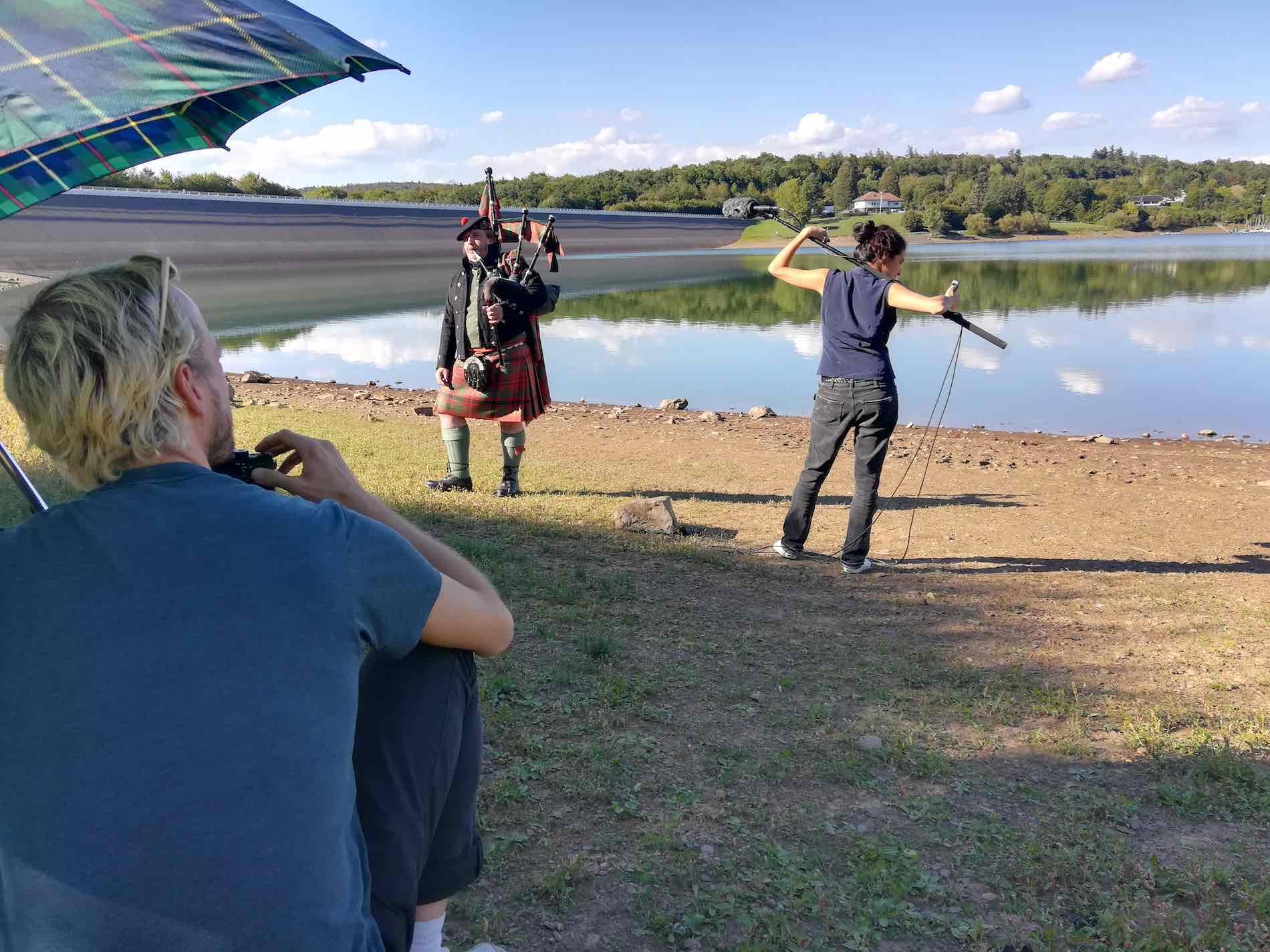 A person in folkloristic Scottish dress and bagpipes stands in front of a dam. Next to it stands a person with a clay rod and in the foreground, under a parasol, a person kneels at a camera.