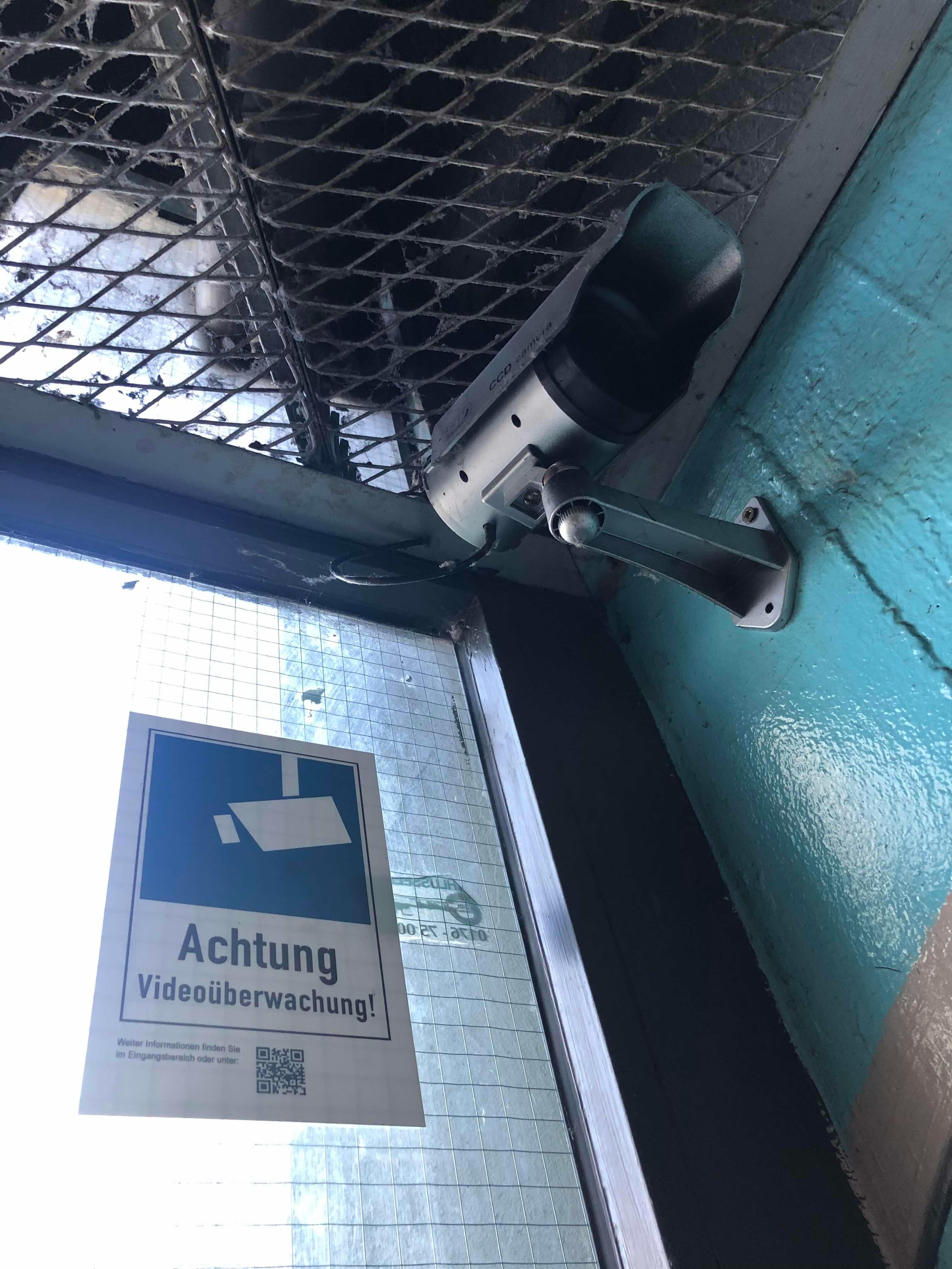 A surveillance camera mounted on the ceiling with a metal grid. Next to it, there is a sign that says 'Warning: Video Surveillance!' with a QR code for more information.