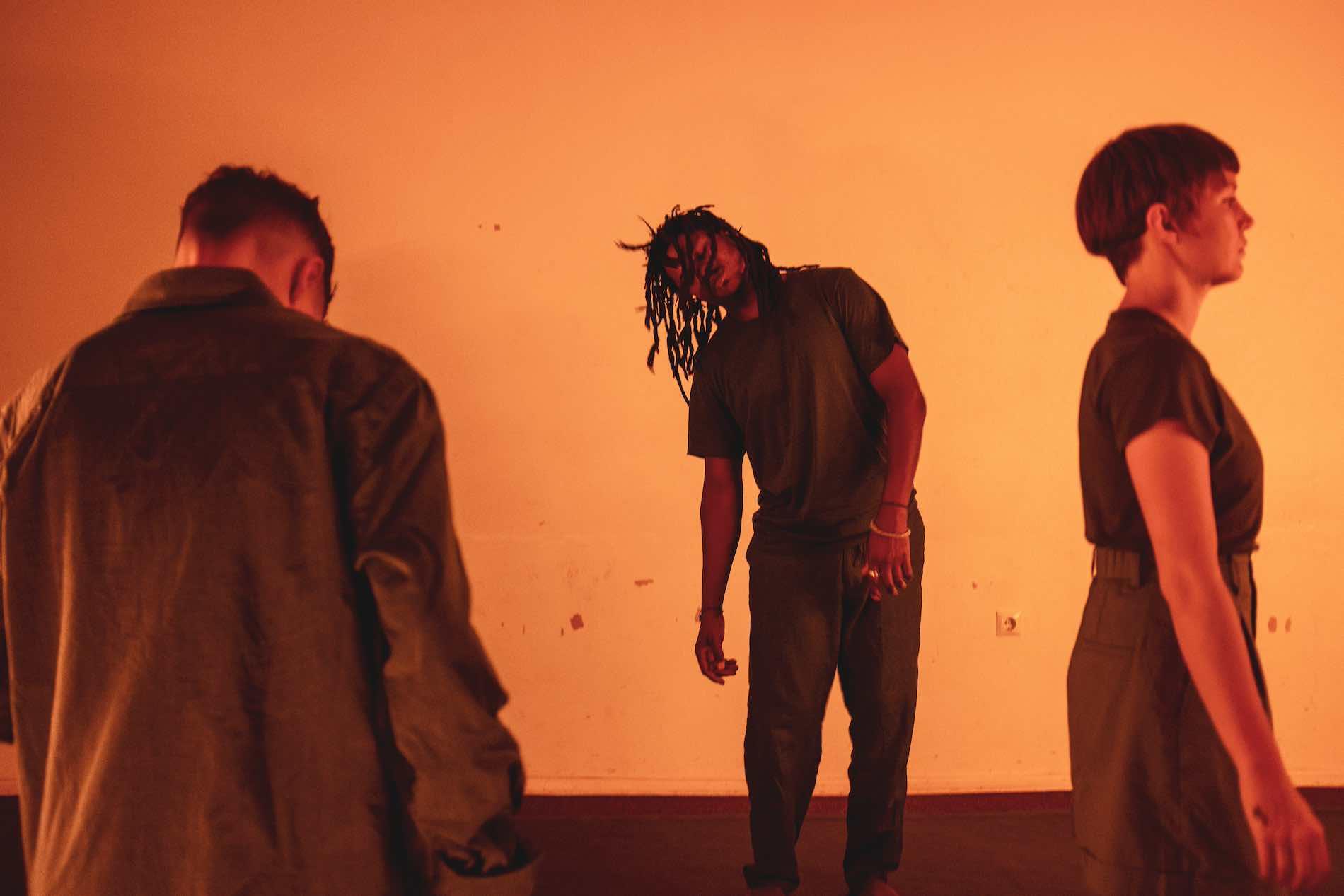 Bathed in orange light, three performers stand in the room, looking in different directions and appearing to be controlled by others.