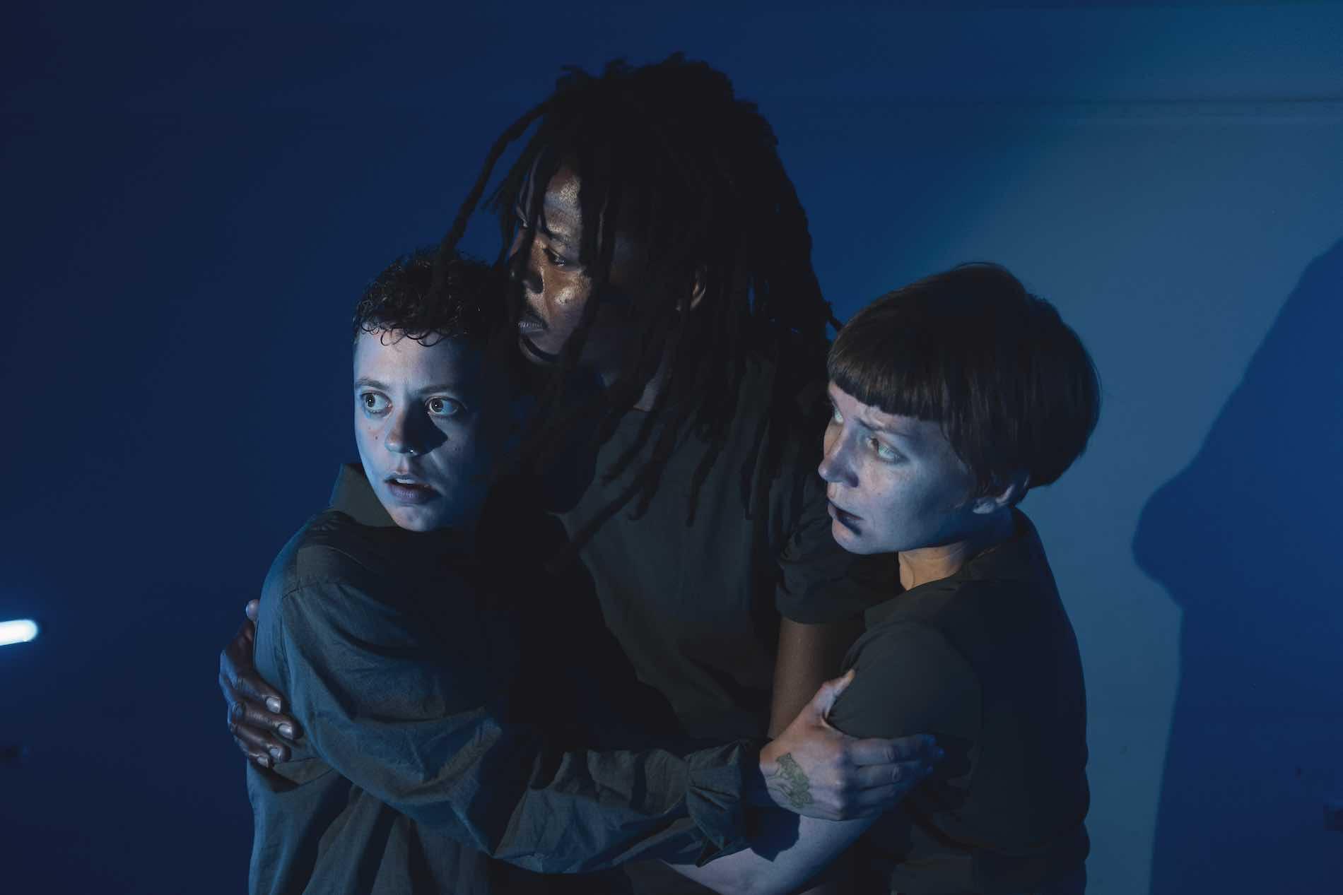 3 people hold on to each other in a half-embrace. The blue light draws attention to their frightened faces.